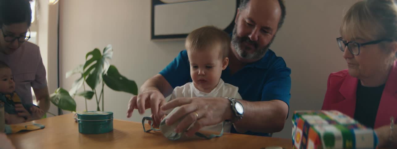 Family sitting at table watching a child with the help of the father opening a gift