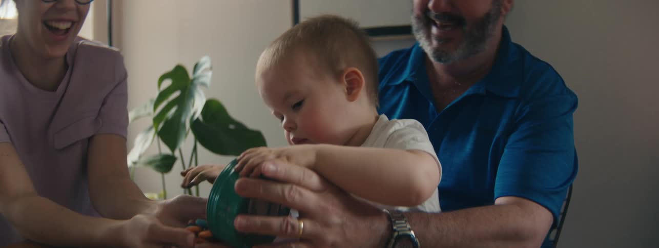 Baby sits in the lap of their grandfather while emptying a container into their mothers hands