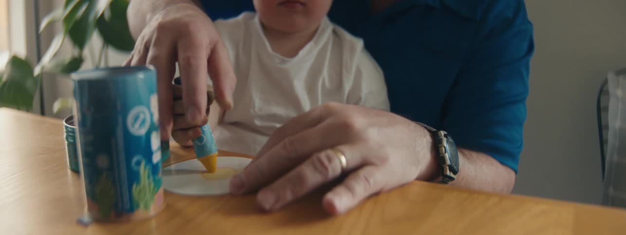 Little boy sitting on grandfathers lap playing with crayons on the table