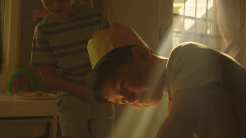 Friends watch a paper crown wearing boy while they play in a shadowy room