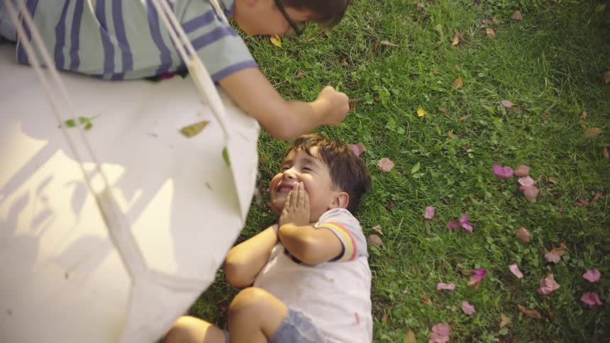 A boy lying on a large swing passes over another boy lying on the ground laughing