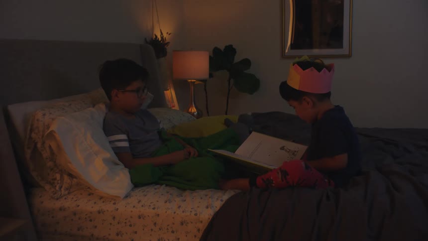 Two boys sit in bed and read a book together in a dimly lit room