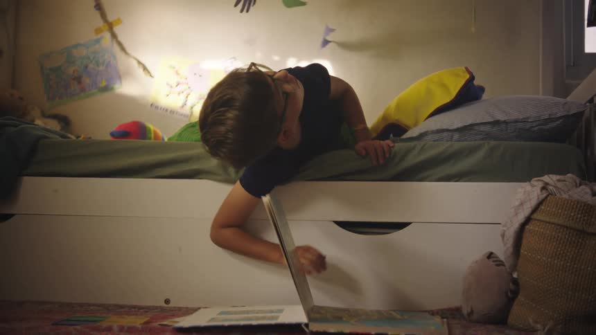 A young boy is lying on his bed reading a book turns away letting the pages fall