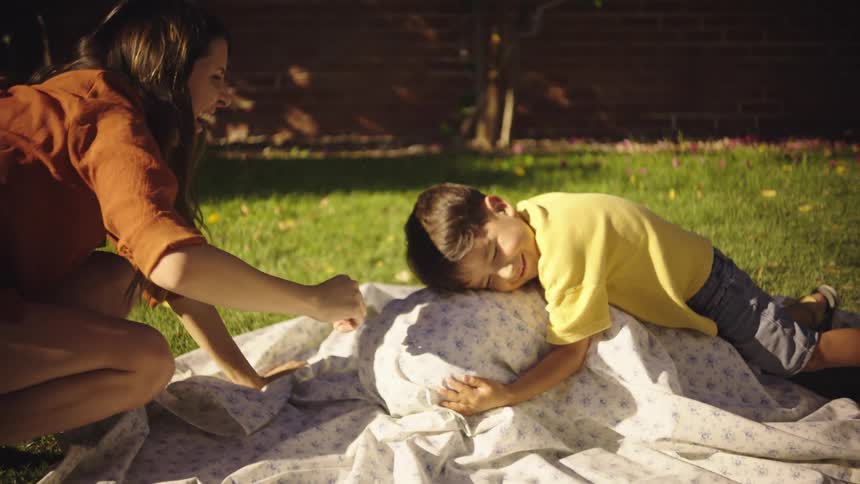 A woman smiles as she plays with two children in a backyard with one of them lying on the grass wrapped in a sheet and the other hugs him laughing