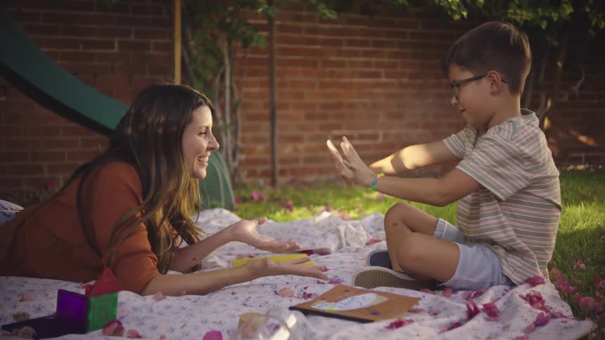 A son makes his mother laugh by demonstrating a magic trick outdoors while the two lounge on a blanket in a yard