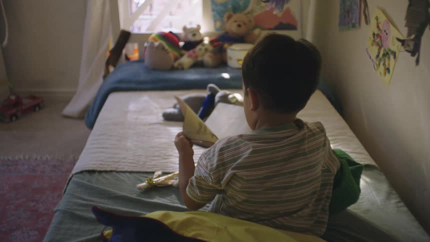 A young boy in bed picks up a book and starts turning pages
