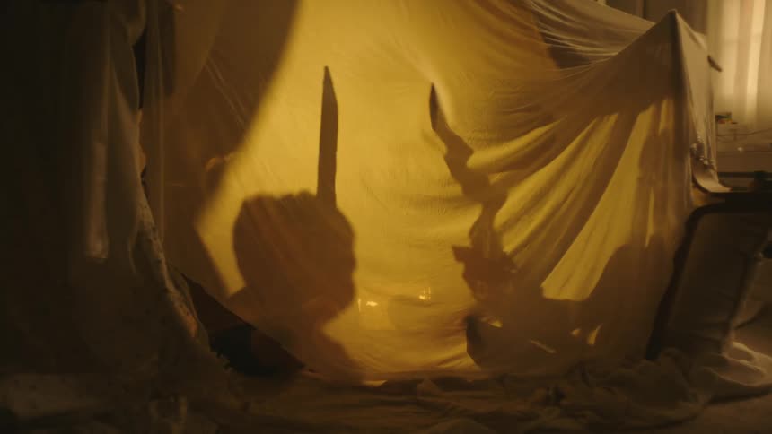 Two children are playing with toy swords inside a homemade tent indoors