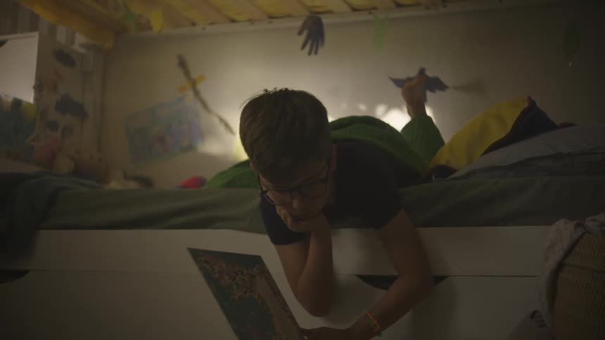 A young boy lies in bed reading while he kicks his feet and lower leg up and down