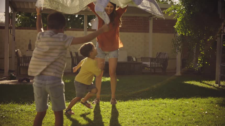 Mother and son lifting a blanket and the other small child runs underneath in the backyard