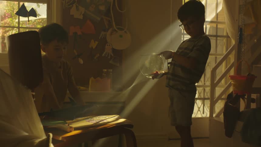 Two brothers in a bedroom attempt to set paper on fire by using a sunlight magnified through a glass fish tank