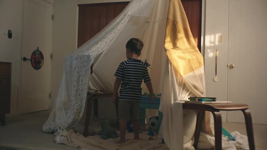 Little boy entering a room closing the door and heading towards a sheet hut with toys