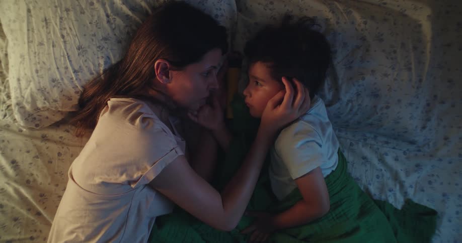 A young mother caresses her young male child while they lie in bed together