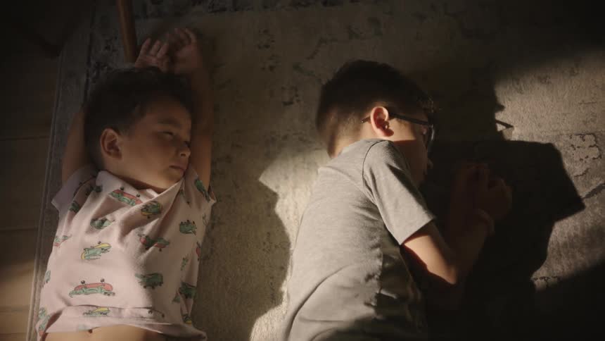 Two children lying next to each other on a floor carpet with one sleeping soundly while the other stretches then flinches when sunlight crosses his face