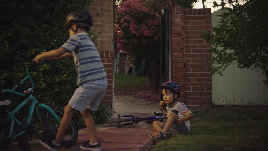 A brother gets off his bike and helps his brother who fell off his bike in a yard