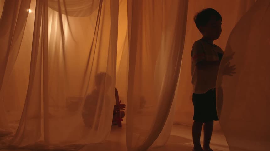 Two boys surrounded by billowing fabric in a lighted room where one stands and the other plays with a fake sword while sitting on the ground