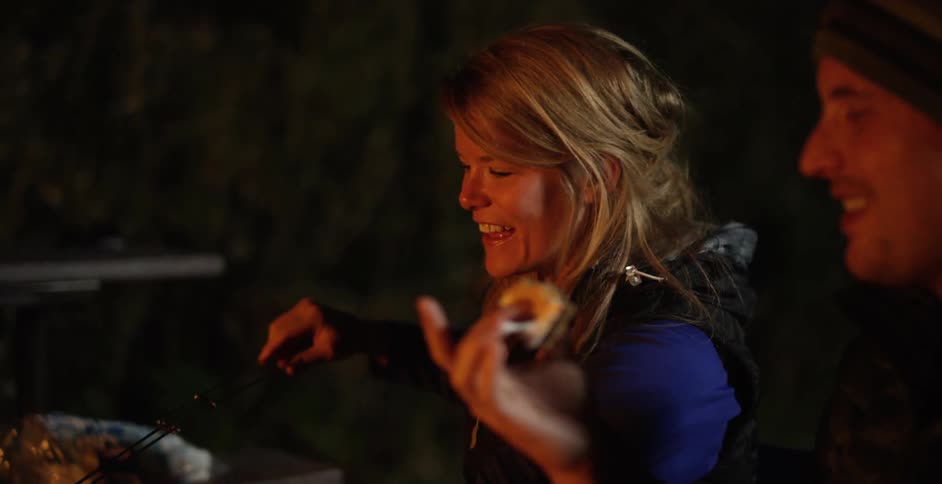 A man holds a piece of food, while a woman stirs something in the dim light