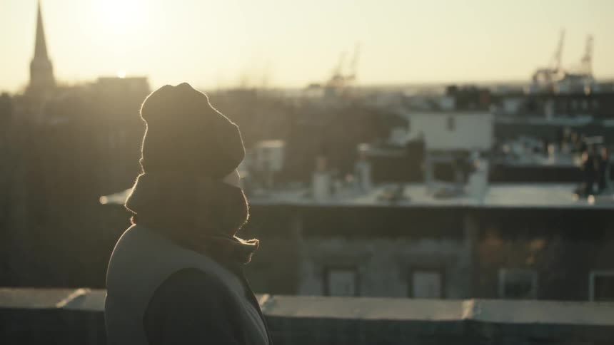 Person standing rooftop in a cap and jacket looking over a city at sunset