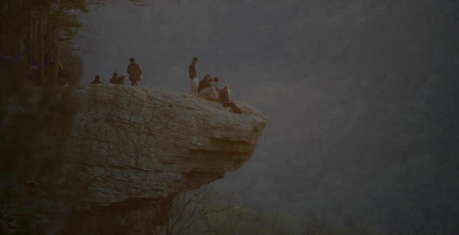 Many people sit and stand on a cliff overlooking a wooded valley