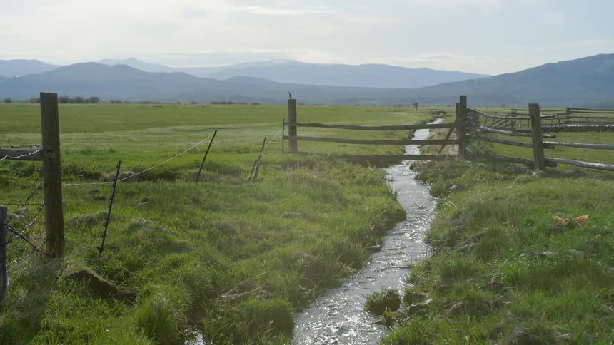 Landscape of a narrow river flowing through a farm with mountains in the background