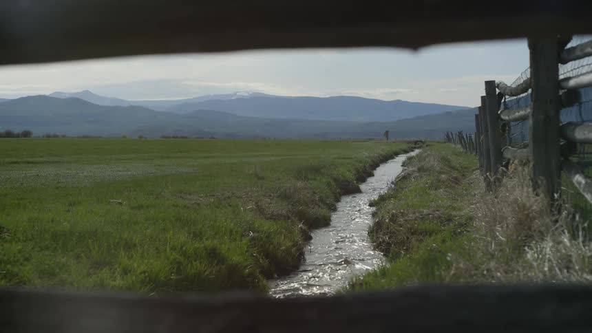 Narrow stream flows through a field next to a wooden fence on a hazy day