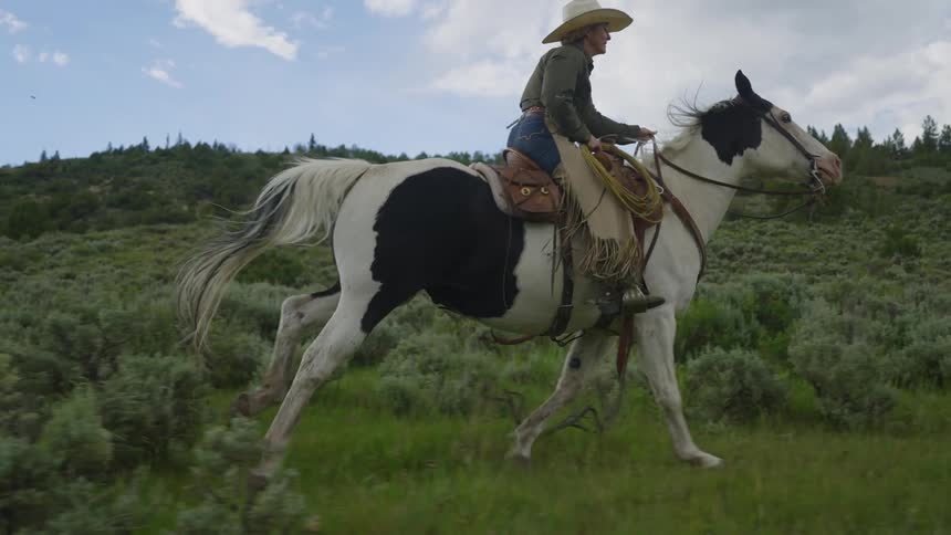A woman wearing a hat is riding a black and white horse quickly in a grass field