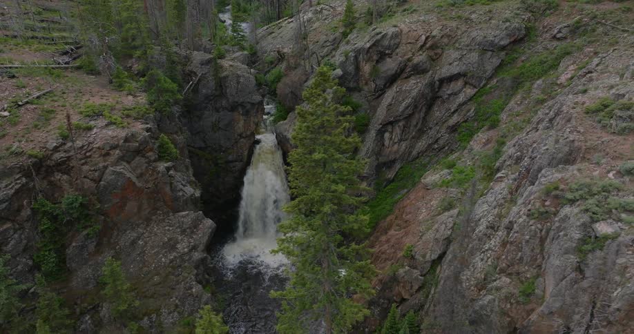 A waterfall cascading down a rocky cliff into a pool below surrounded by pine trees and rugged terrain