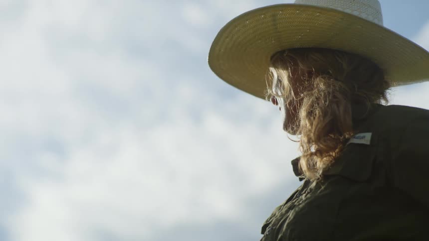 A woman in a coat and wearing a straw hat looks off into the distance outdoors