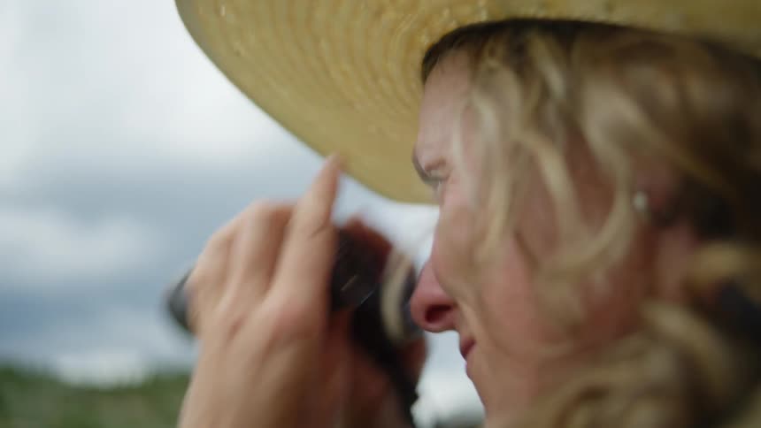 A blonde female wearing a straw hat looks into the distance through binoculars and then smiles