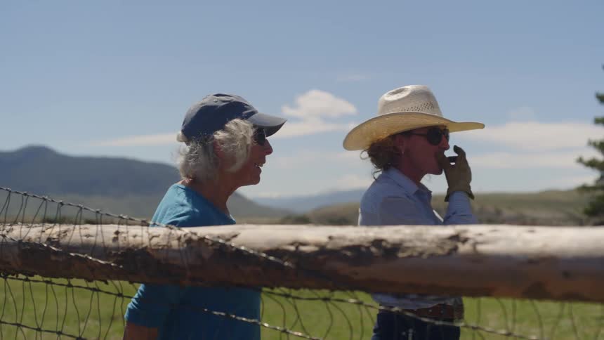 Two women standing by a wooden fence with a vast grassy field and mountains in the background