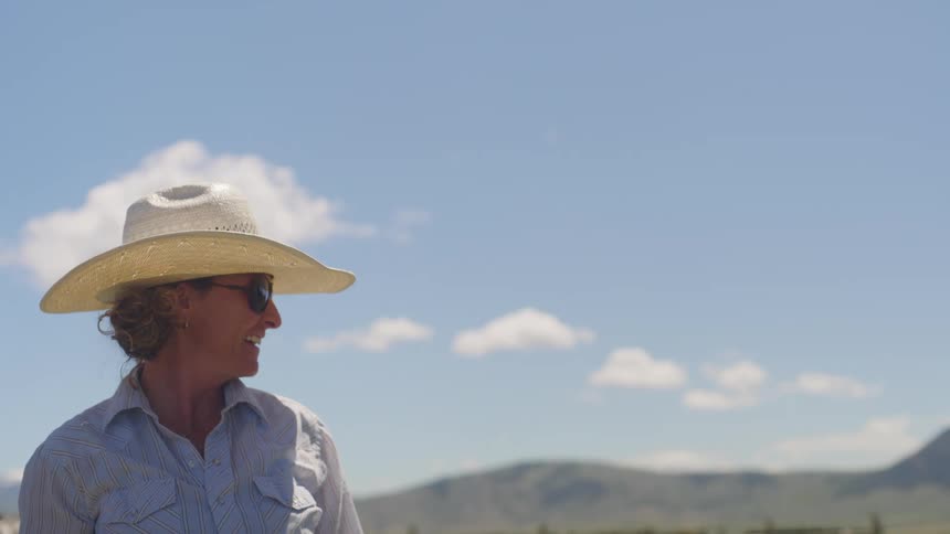 Woman wearing sunglasses and a hat pointing in a direction with mountains in the background