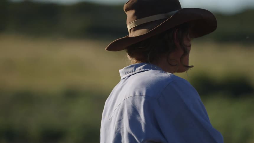 A woman wearing a widebrimmed hat and a light blue shirt rides a horse