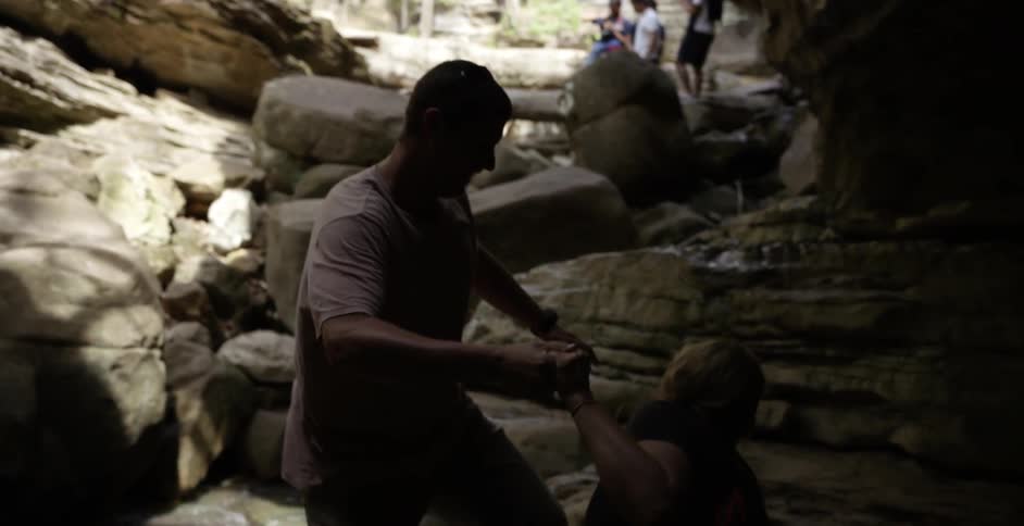 A woman holds a man's hand as she jumps down from a small cliff and laughs