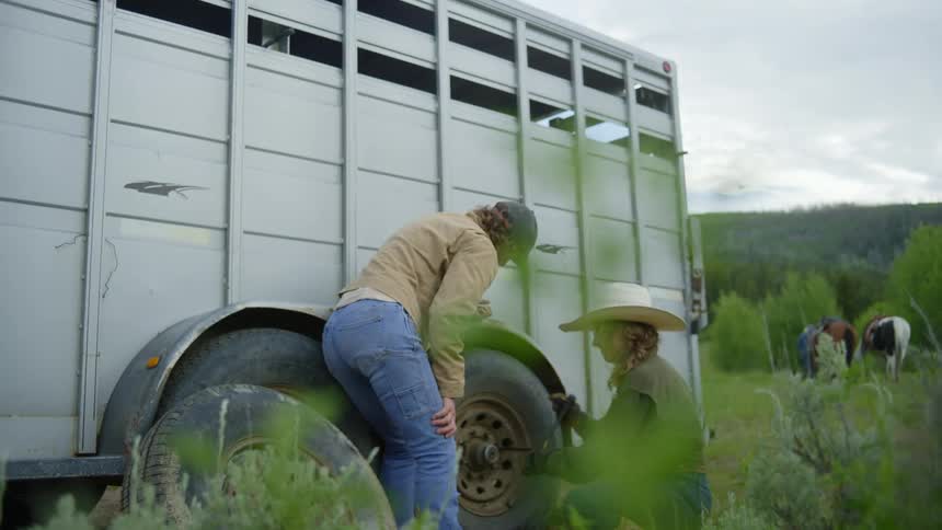 A person bends over the wheel of a large white trailer to change it in a rural area