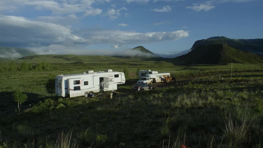 A trailer and some cars parked in a grass field and there are some horses next to it