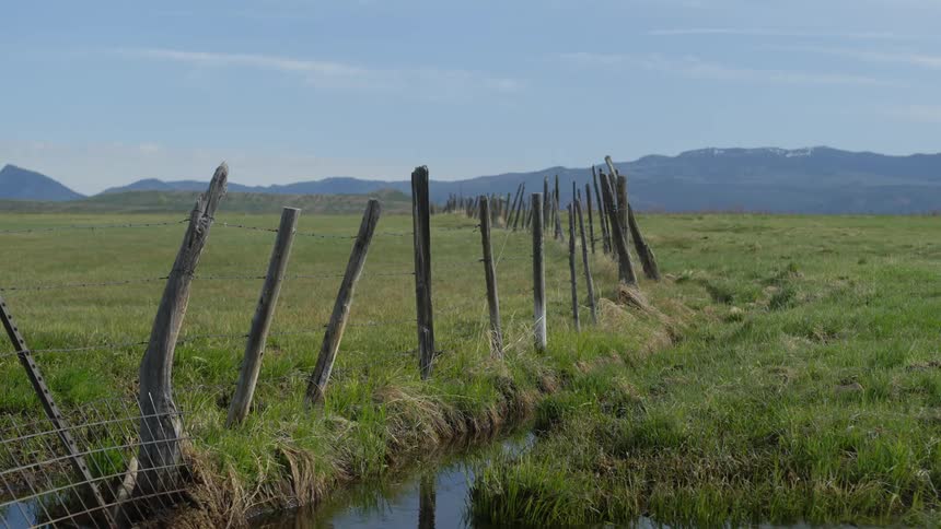 Narrow river next to a wooden fence with barbed wire in a pasture and mountains in the background