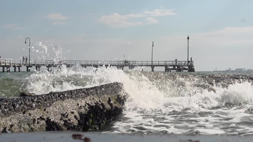 Waves crashing off rocks near a pier on a cloudy day