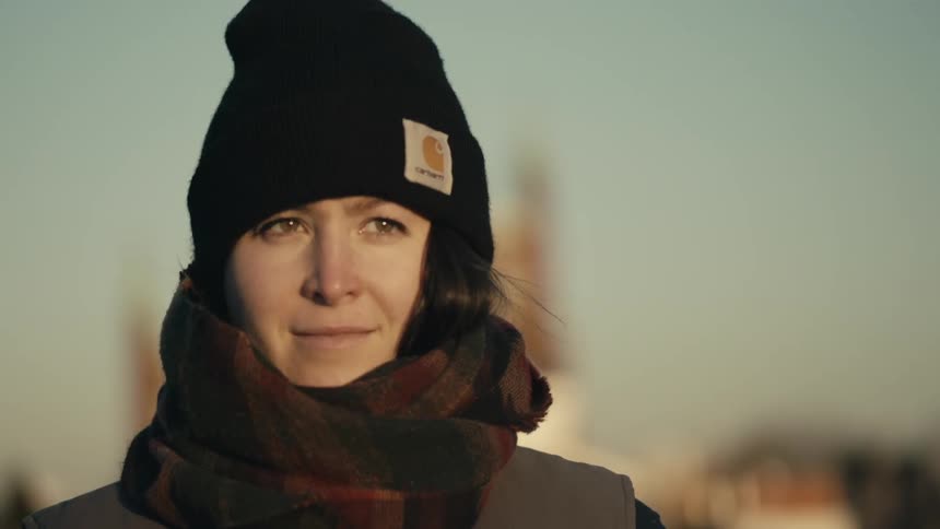 A woman wearing a knit cap and winter scarf stands outside and smiles