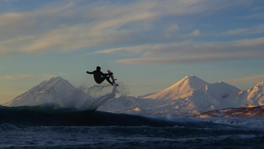 Surfer flying above a wave with a board and snowy mountains on the horizon