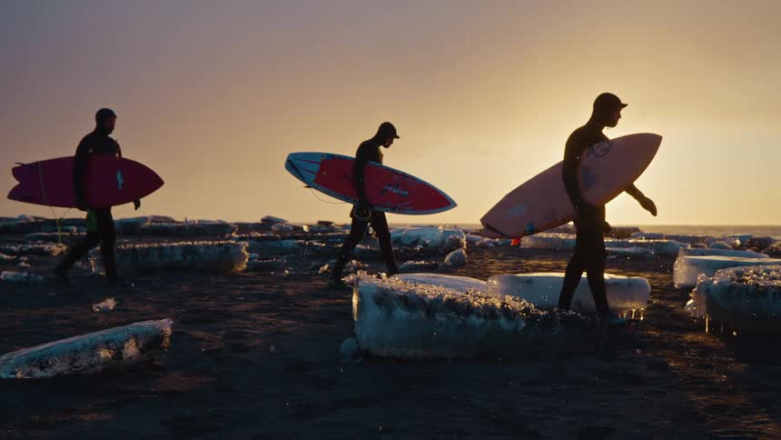 Three men with surfboards walking between blocks of ice at sunrise