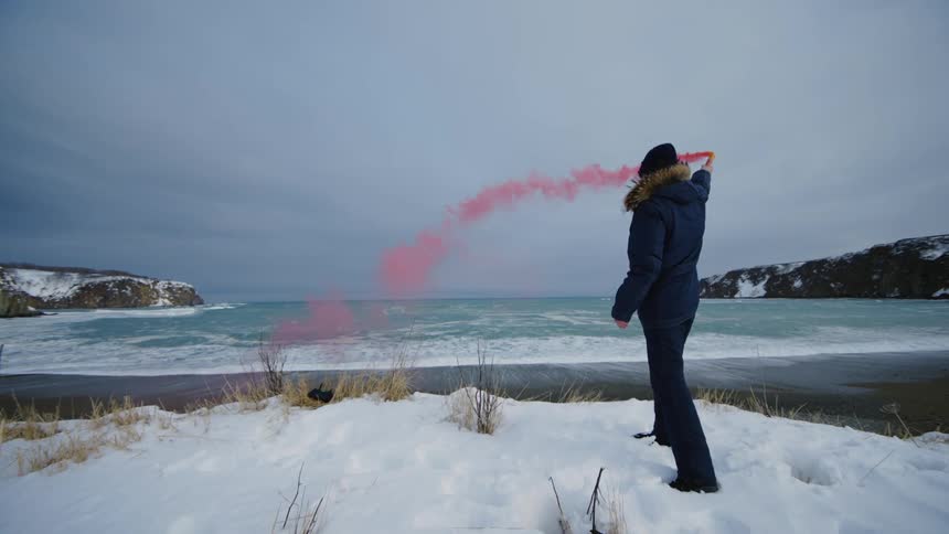A person stands on a snowy shore facing the sea holding a red smoke flare