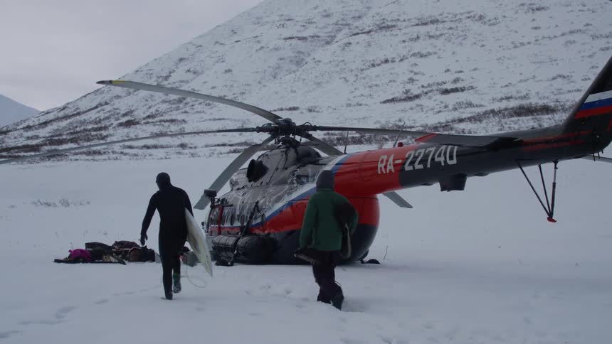 Two people walking towards a helicopter that is landing in a snowcovered environment on the edge of a mountain