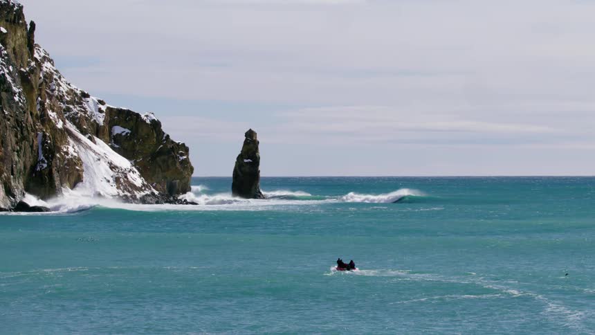 A boat with people moves to an extremely rocky coastline partially covered in snow