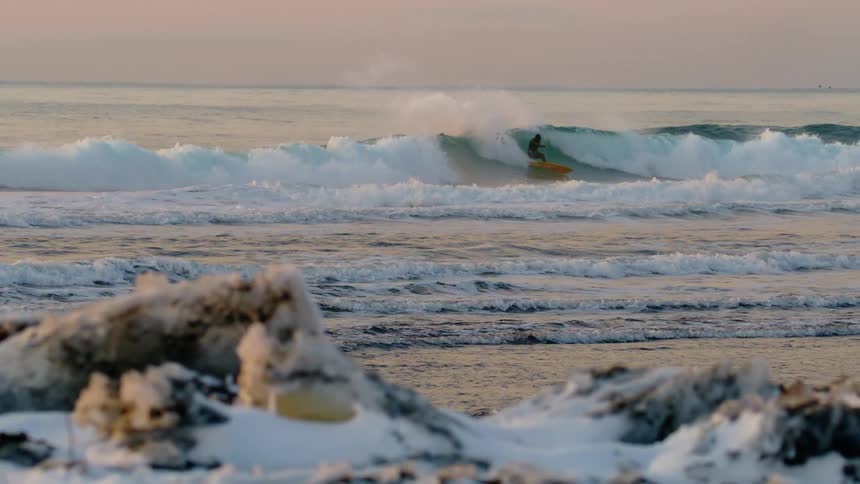 Seascape with snow in the foreground and a person surfing a wave in the background