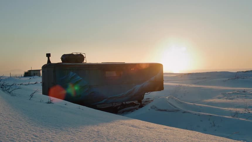 A camper sits on dirt and is surrounded by snow as the sun sits on the horizon
