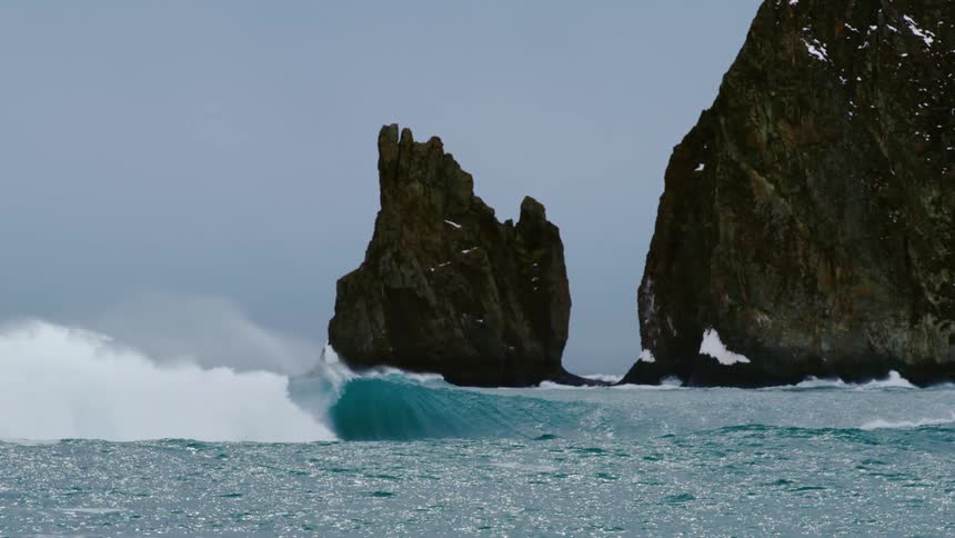 Strong ocean wave crashing against the rocky snowy coastline in the background
