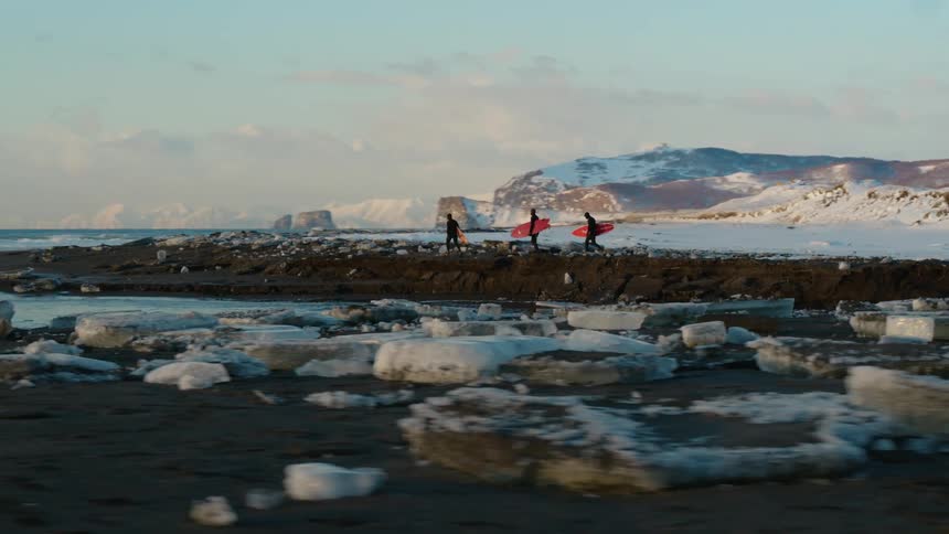 Three people with surfboards walking on snowy and icy terrain towards the sea