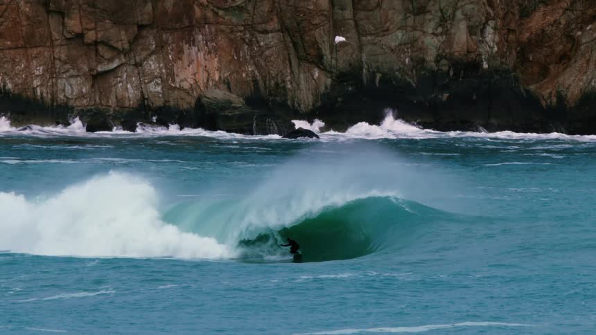 A surfer rides a breaking wave with rocky cliffs in the background
