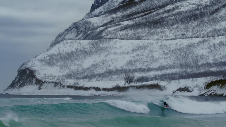 Lone surfer doing tricks on a wave with the coast in the background covered in snow