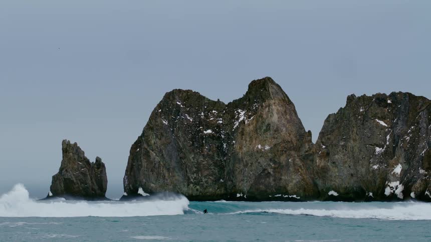 A surfer rides waves near a large rugged rock formation