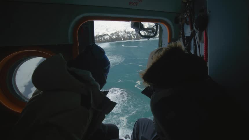 Two people looking out to sea while in the cockpit of a helicopter flying overhead